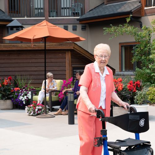 A female senior resident with a coral outfit uses a black and blue walker as she strolls through a courtyard in front of two other female residents sitting in lounge chairs.