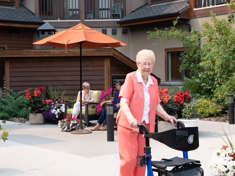 A female senior resident with a coral outfit uses a black and blue walker as she strolls through a courtyard in front of two other female residents sitting in lounge chairs.