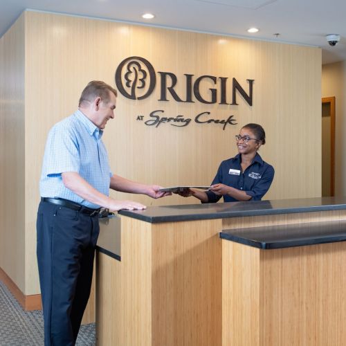 A smiling female staff member in a black uniform hands a male senior resident a form at the Origin at Spring Creek front desk.