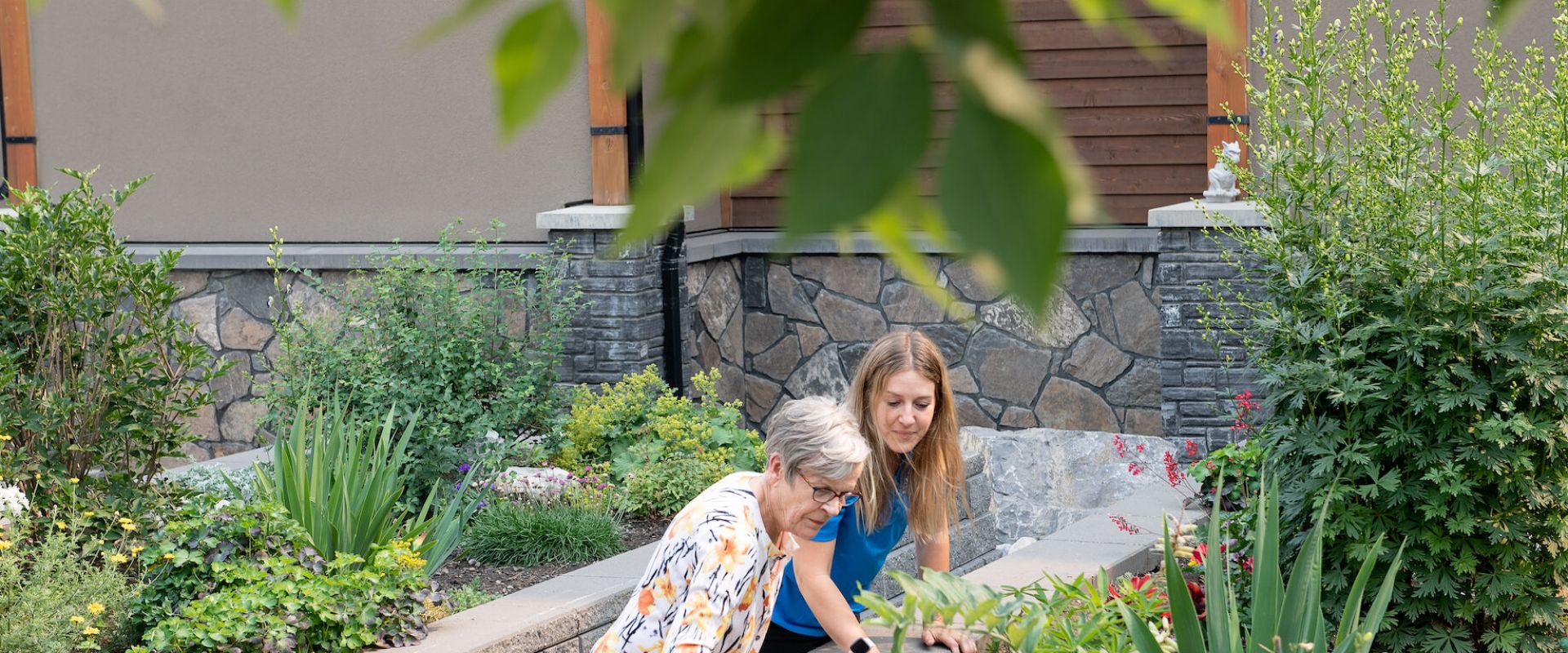 A female senior resident at Origin at Spring Creek and a Life Enrichment Coordinator in a blue uniform look at flowers and greenery in a garden space at Spring Creek