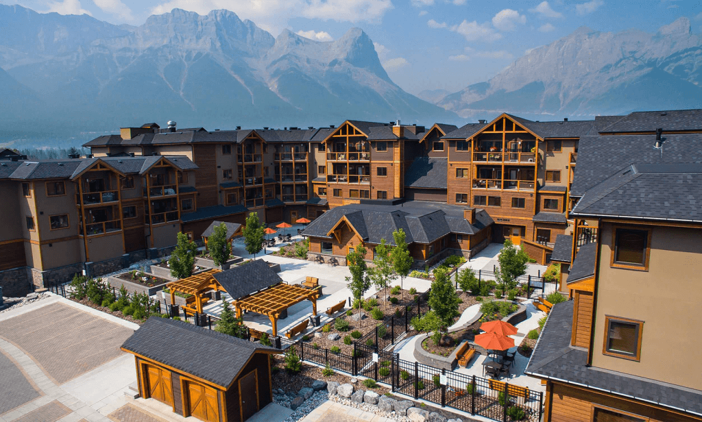 An above view of the beautiful living complex at Origin at Spring Creek on a sunny day, with a lush courtyard full of green plants and comfortable seating, warm paint colours and the gorgeous Rocky Mountains in the background in Canmore, Alberta.