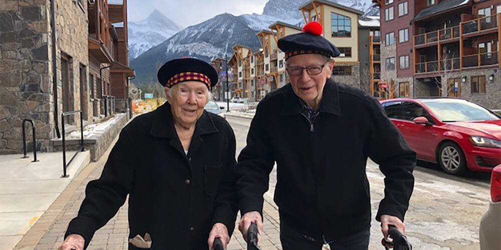 A senior woman and man walk down the street in Spring Creek Mountain Village in Canmore, Alberta, with the mountains in the background. 