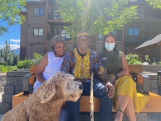 Three people are sitting outside of the building two Origin residents Jane and Richard, and Life Enrichment Educator Zoe Robinson. Additionally, a dog is sitting beside them.