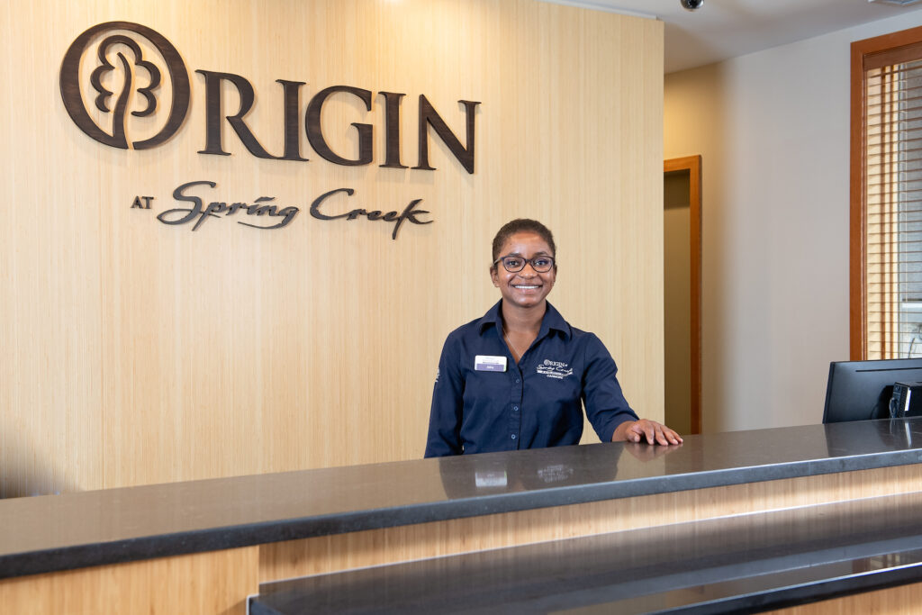 A female Origin at Spring Creek front desk team member standing behind the front desk at Origin at Spring Creek, smiling and welcoming you to the facility.