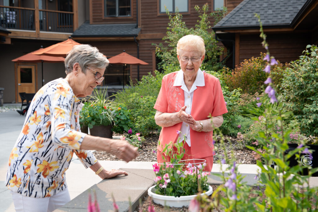 Two female senior residents stand in the courtyard by a lush garden bed, tending to a white pot of pink and white flowers.