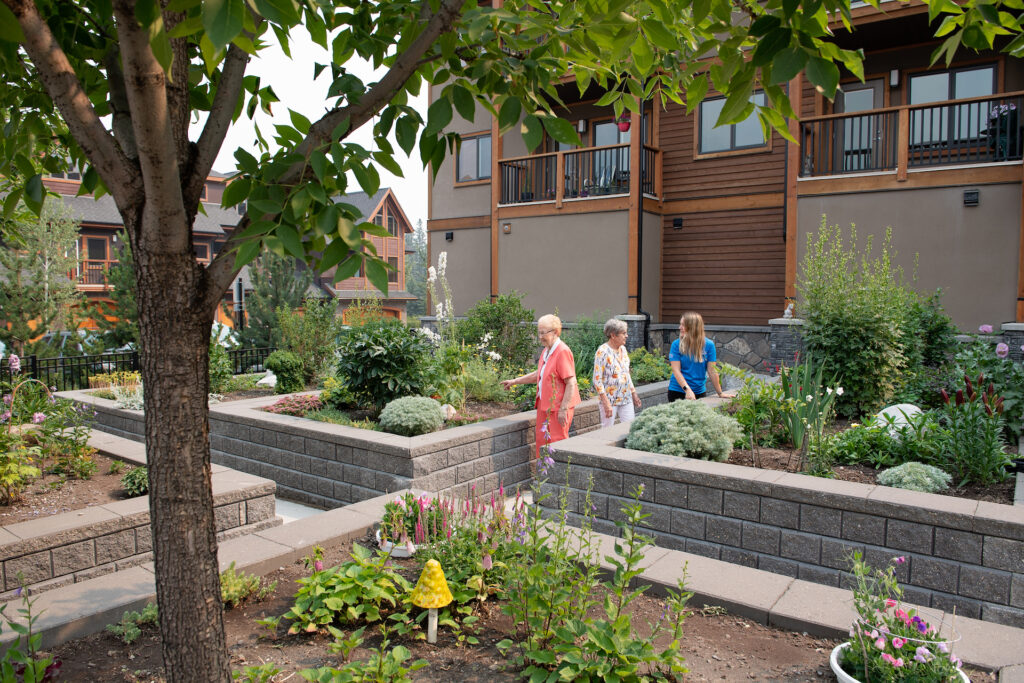 Two female senior residents and a female Life Enrichment Coordinator walk beside the brick-lined garden beds together, looking at the foliage, lush flowers and a large tree nearby.