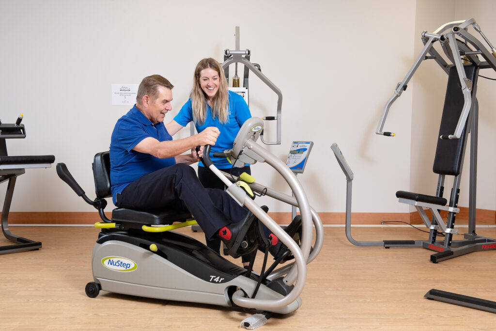 A smiling female Origin companion in a blue uniform assists a senior resident as they use an elliptical exercise machine in the facility's gym.