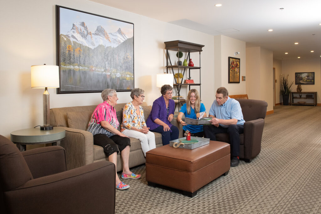 Four senior residents and a female Life Enrichment coach look over a magazine as they sit on a comfortable couch in one of Origin at Spring Creek's living spaces.