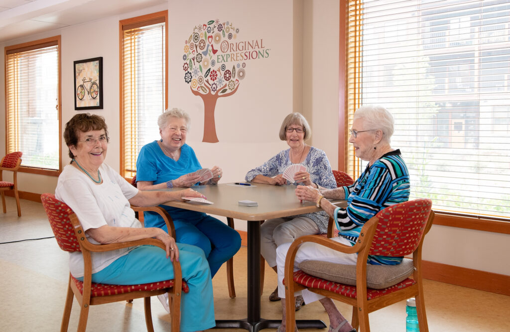 Four smiling female senior residents all dressed in blue and white enjoy a game of cards as they sit at a table in front of a bright, sunlit window.