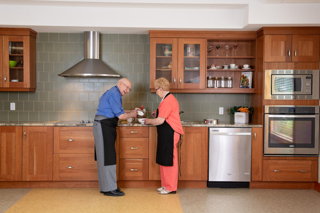 A male senior resident in a blue shirt and a female senior resident in a coral outfit cook together in a large, open kitchen with grey-green tiles, wooden cabinets, and stainless steel appliances.