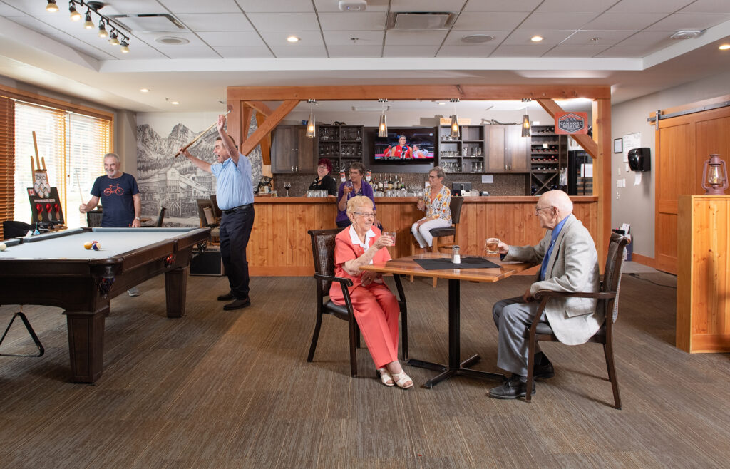 Six senior residents enjoy their time in the Origin at Spring Creek amenities room, where a female Origin staff member in a black uniform serves drinks from the bar, with two of the male senior residents playing a game of pool to the left, two female residents drinking wine at the bar, and another two senior residents sip on drinks at a table in the front.