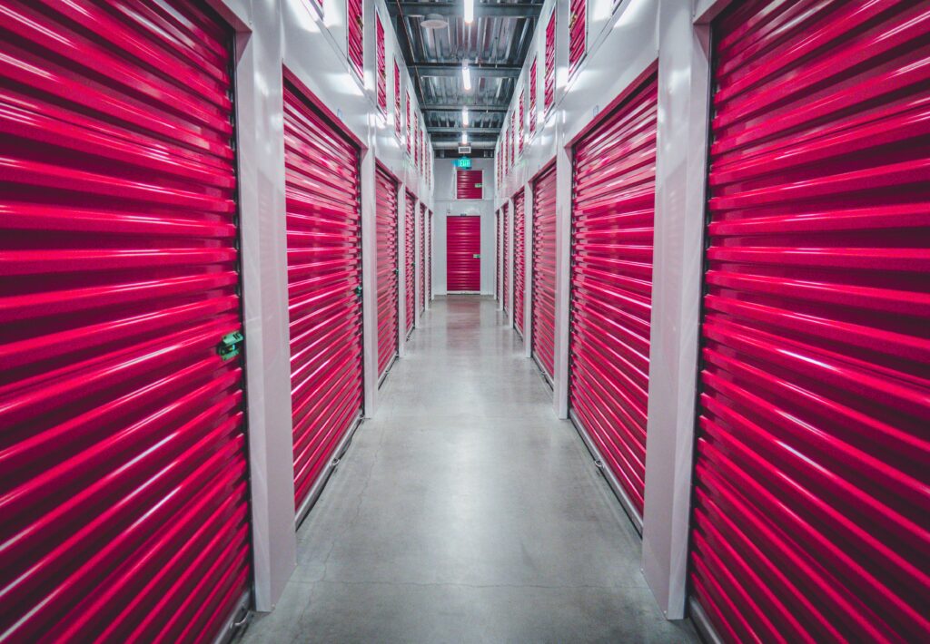 An aisle of bright red storage unit in a grey cement hallway stretches out in a rental storage facility.